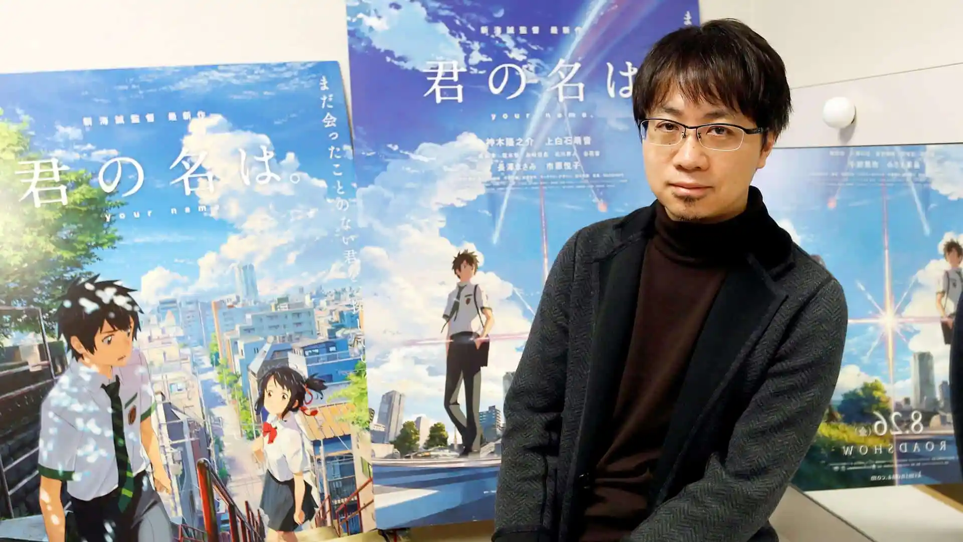 Makoto Shinkai sorrindo em frente a um pôster de "Your Name".